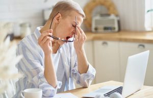 Tired sleepy middle aged woman massaging her nose and yawning while working on laptop online from home, sitting in the modern kitchen at home. Senior people and distance work concept